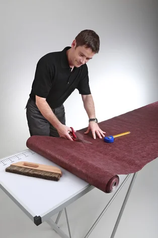 Illustration - Ihr Experte für schönes Wohnen - Ein Handwerker schneidet eine Rolle Tapete auf einem Tapeziertisch